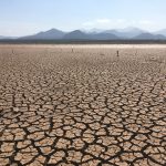 Paisagem rural extensa com o solo severamente rachado sob céu aberto, indicando condições de seca.