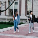 Grupo de estudantes caminhando por uma calçada no campus universitário, alguns carregando mochilas.