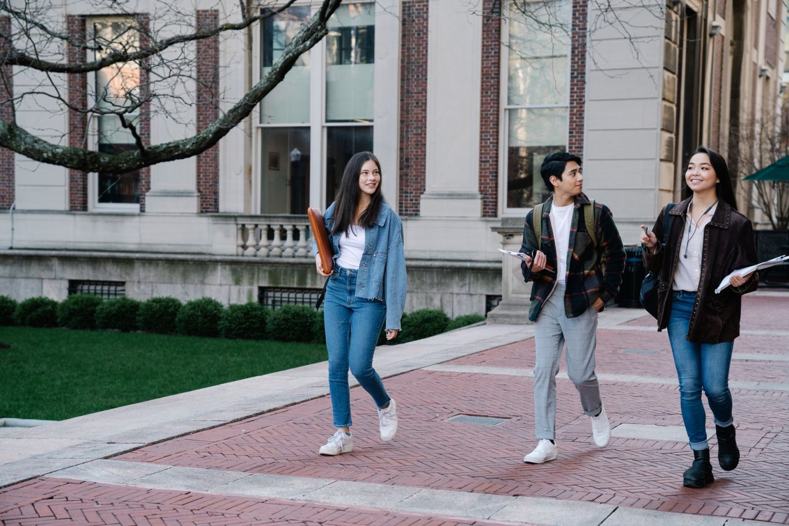 Grupo de estudantes caminhando por uma calçada no campus universitário, alguns carregando mochilas.