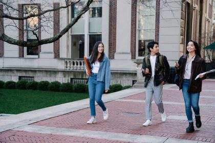 Grupo de estudantes caminhando por uma calçada no campus universitário, alguns carregando mochilas.