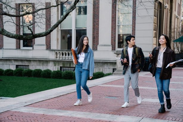 Grupo de estudantes caminhando por uma calçada no campus universitário, alguns carregando mochilas.