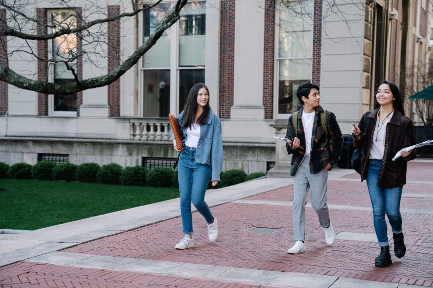 Grupo de estudantes caminhando por uma calçada no campus universitário, alguns carregando mochilas.