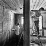 Dois trabalhadores reformam o interior de uma casa; um deles aplica reboco enquanto está sobre uma escada.