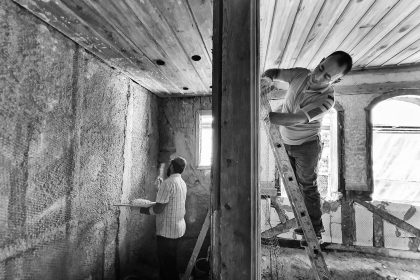 Dois trabalhadores reformam o interior de uma casa; um deles aplica reboco enquanto está sobre uma escada.