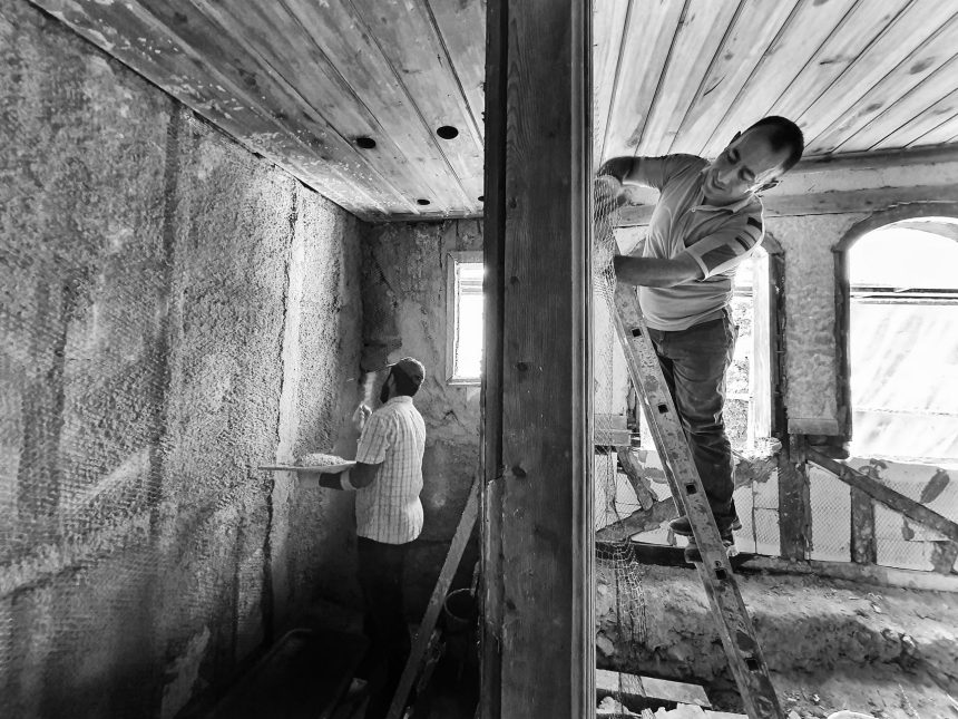 Dois trabalhadores reformam o interior de uma casa; um deles aplica reboco enquanto está sobre uma escada.