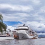 Navios de cruzeiro atracados em um porto tropical, com palmeiras ao redor e céu nublado.