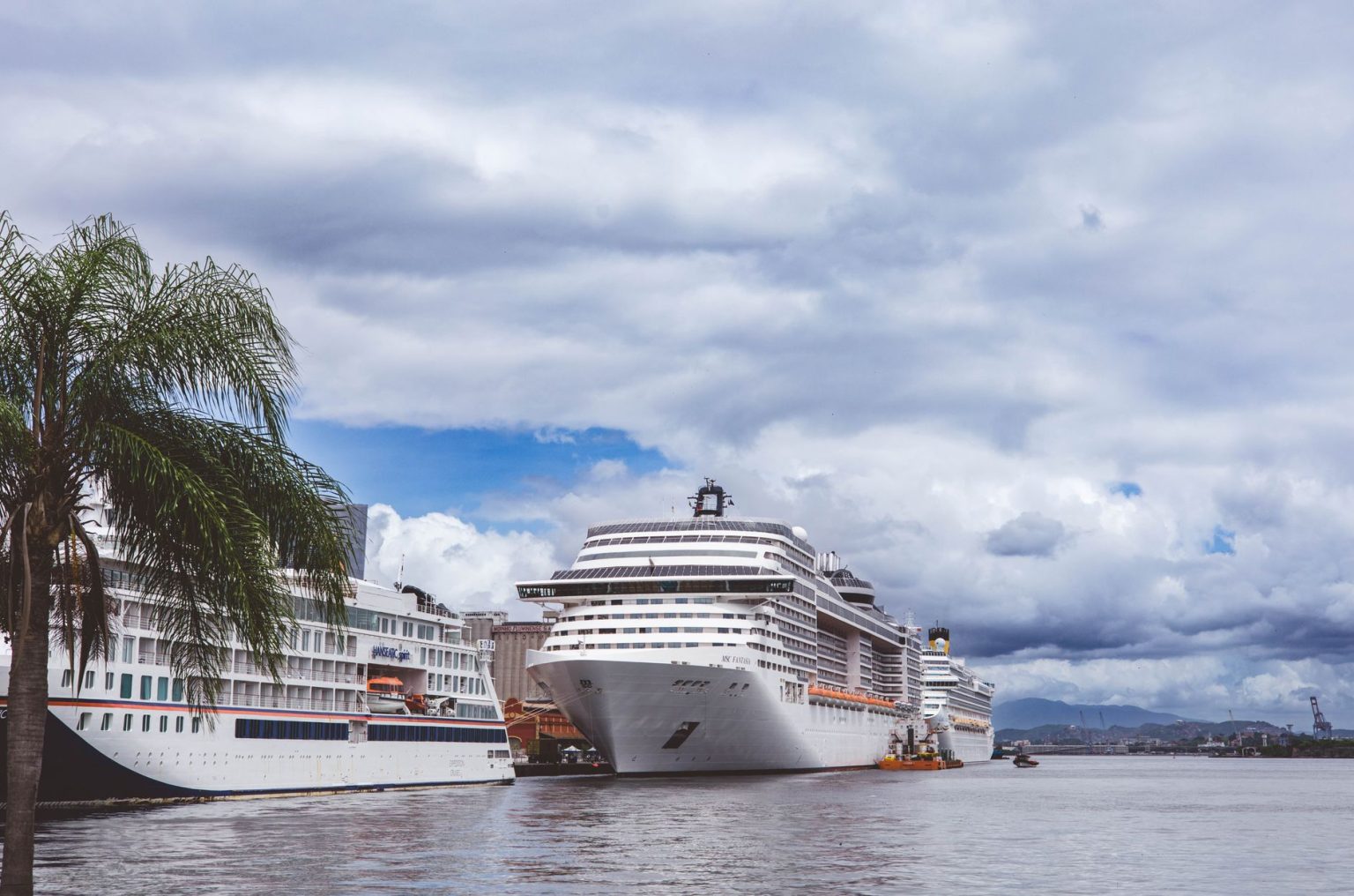 Navios de cruzeiro atracados em um porto tropical, com palmeiras ao redor e céu nublado.